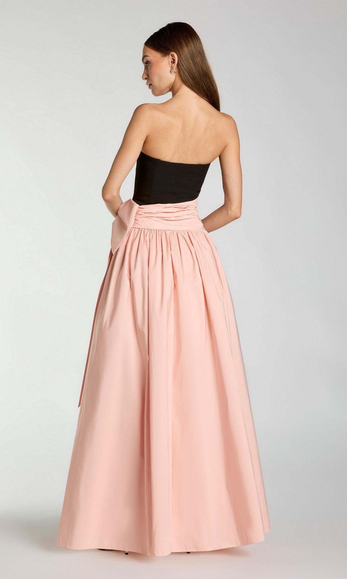 A woman stands with her back to the camera against a plain background, wearing the Long Formal Dress: Mac Duggal 2370, which features a strapless black bodice and a long, full pale pink skirt.